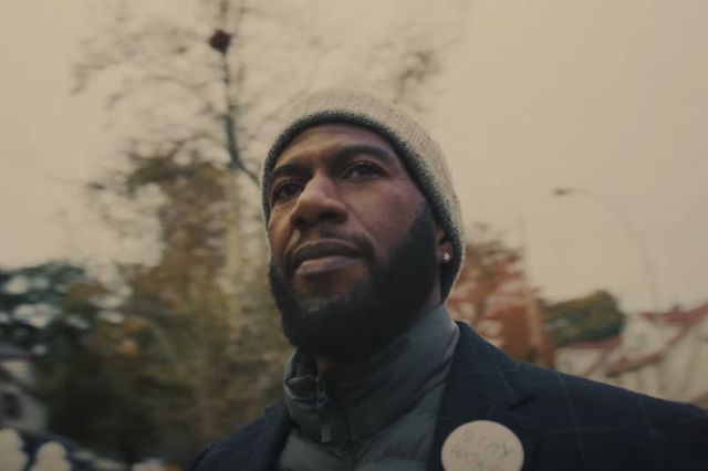 Jumaane Williams in a knit cap and coat looking at a house while on a sidewalk